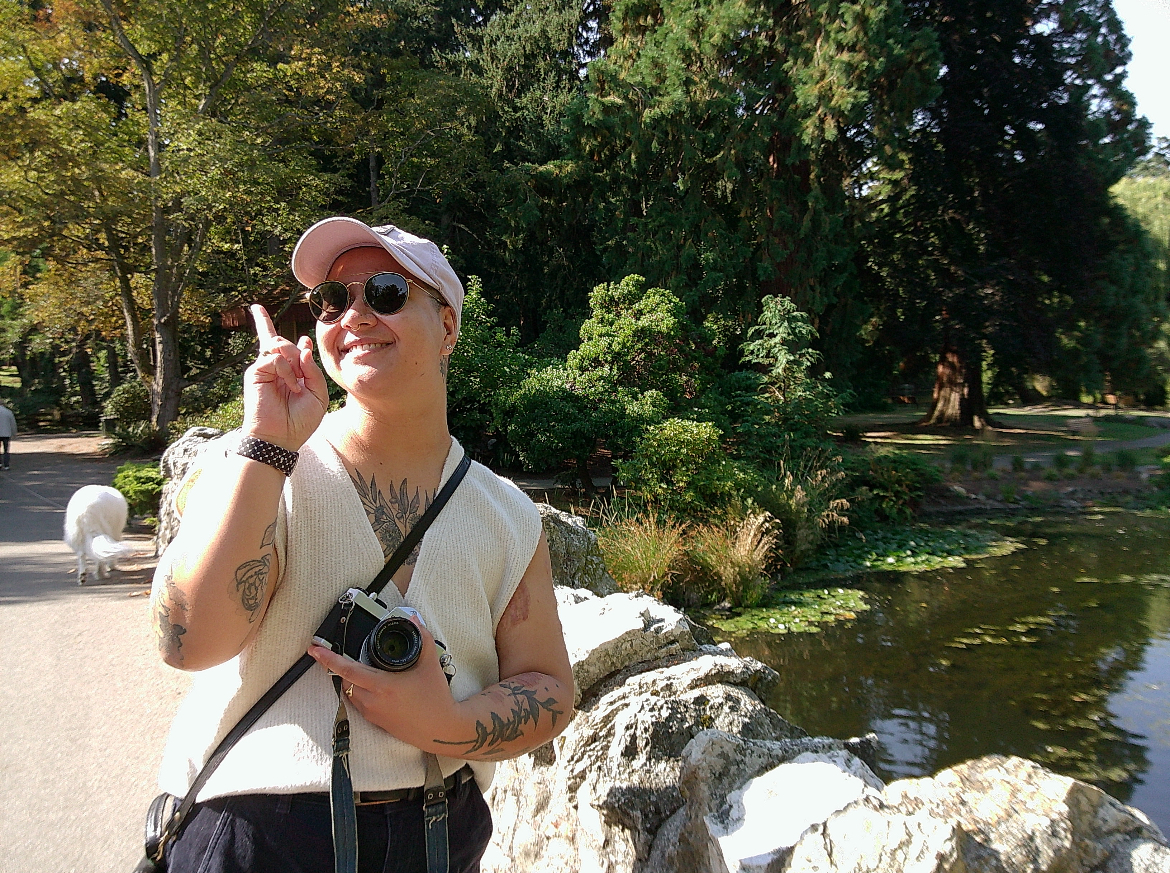 Maria posing in front of greenery and a water feature. Maria is wearing a white sweater vest, purple ball cap, and sunglasses. They are holding a film camera.