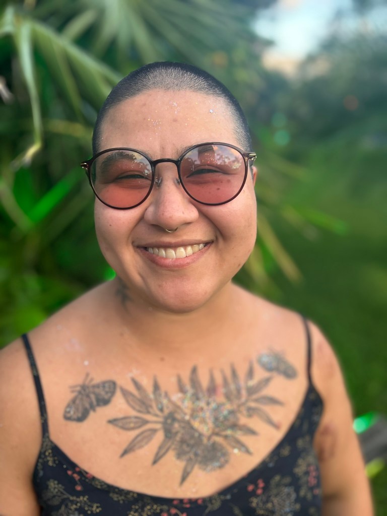 Portrait of Maria smiling in front blurred greenery. Maria is wearing a dark blue tank top, revealing a botanical chest tattoo. Maria is wearing lightly shaded, round framed glasses.